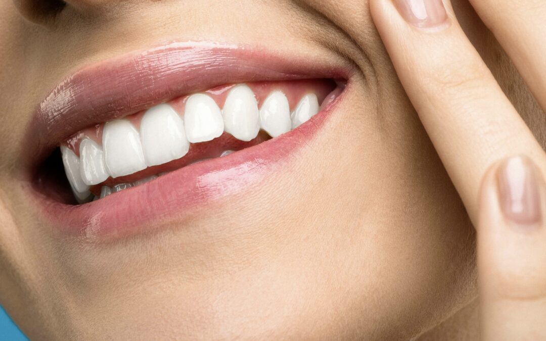 Close-up of a smiling person’s mouth with glossy pink lips, straight white teeth made with cosmetic veneers in midlothian va, and fingers gently touching the cheek.